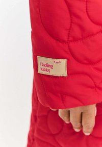 Czerwona pikowana rękaw kurtki z fakturowaną powierzchnią oraz beżową metką z napisem "Feeling lucky" w kolorze różowym.