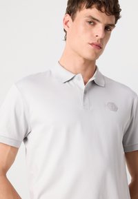 Polo cinza claro feito de tecido suave, com colarinho, mangas curtas e um logótipo hexagonal subtil no lado esquerdo do peito.