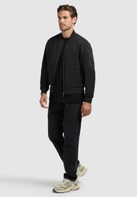 Homme debout portant une veste bomber noire, un pantalon noir et des baskets beige sur un fond clair uni.