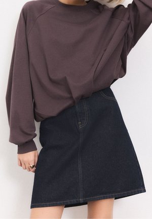 Person iført en løs mørkelilla langærmet top og et mørkeblåt jeans A-line skørt, med en hånd hvilende nær hoften.
