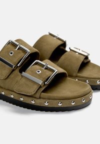 Braune Wildledersandalen mit zwei verstellbaren Riemen, metallischen Schnallen und dekorativen Nieten entlang der Gummisohle. Flaches Design mit runden Zehen.