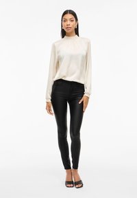 Beige satin långärmad blus med rynkad halsringning, kombinerad med svarta åtsittande byxor och svarta sandaler med öppen tå, visad på en modell.