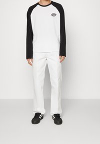 White long-sleeve T-shirt with black raglan sleeves and a logo on the chest, paired with white trousers and black sneakers.