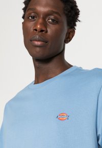 Jovem com cabelo curto e encaracolado, vestindo uma t-shirt azul clara da Dickies, virado para a frente com uma expressão neutra contra um fundo simples.