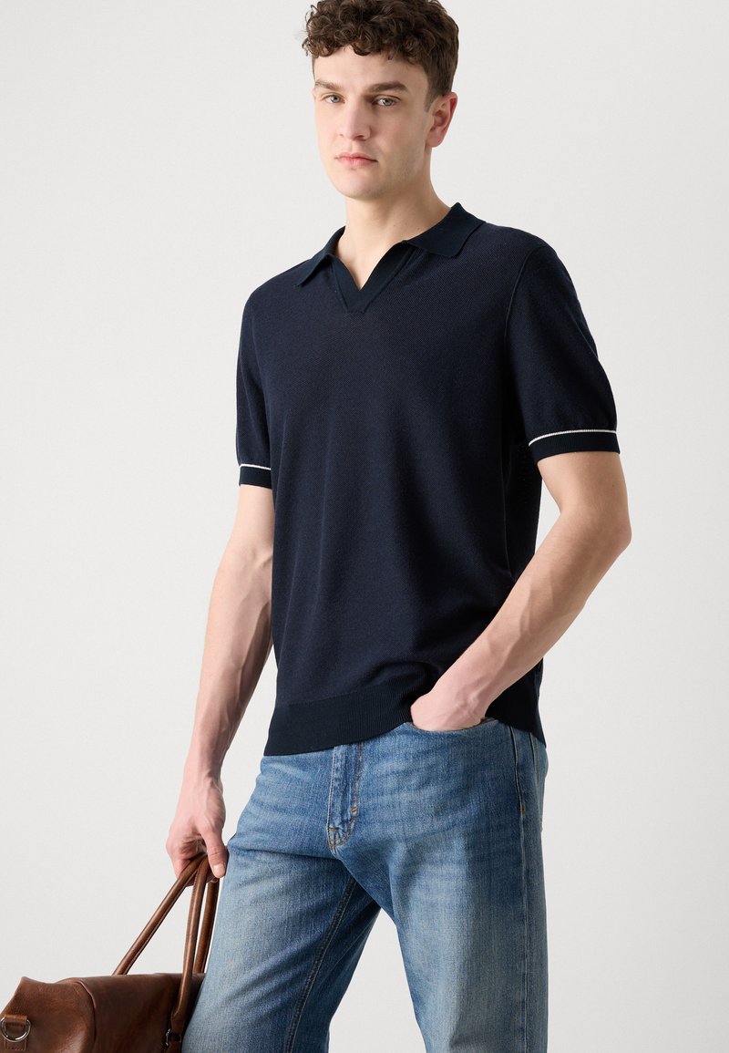 Polo en maille marine à manches courtes, col en V, col contrasté et poignets rayés. Assorti à un jean bleu clair et un sac en cuir marron.