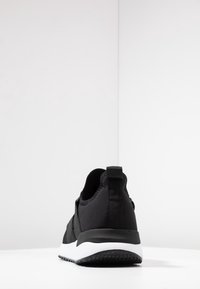 Svart sportssko med överdel i texturerad tyg, dragflik bak, och en kontrasterande vit mellansula med en strukturerad svart yttersula.