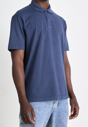 Dunkelblaues Poloshirt aus Baumwolle mit klassischem Kragen, kurzen Ärmeln und einem tonalen Logo auf der Brust. Kombiniert mit hellen Jeans.