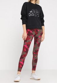 Czarna, długoramienna bluza z dużym, szarym logo "adidas", w połączeniu z dopasowanymi legginsami w kamuflażu w odcieniach burgundowym, różowym i brązowym. Białe sneakersy.