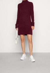 Burgunderfärgad stickad klänning med långa ärmar, hög krage, sidofickor och en strukturerad yta, kombinerad med vita sneakers och en liten väska.