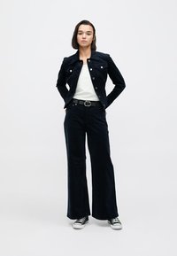 Σκούρο μπλε σκούροι σακάκι από κορδόνι με κούμπωμα με κουμπιά, συνδυασμένο με παντελόνια με φαρδιά μπατζάκια στο ίδιο χρώμα. Κάτω από αυτό, ένα λευκό απλό πουκάμισο και μαύρα αθλητικά παπούτσια.