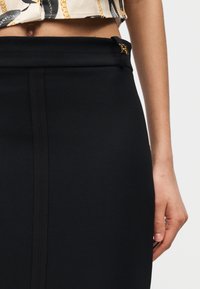 Κοντινή άποψη της μέσης και του χεριού που φοράει μια μαύρη ψηλόμεση φούστα με χρυσή αγκράφα και ένα κοντό τοπ με σχέδια.