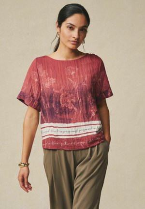 Frau trägt eine rot geblümte, kurzärmelige Bluse, olivgrüne Hose, goldene Creolen und ein goldenes Armband, steht mit einer Hand in der Tasche.