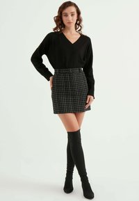 Svart stickad tröja med V-ringning, kombinerad med en tight svart kjol med ett vitt rutmönster. Modellen bär knähöga svarta stövlar.