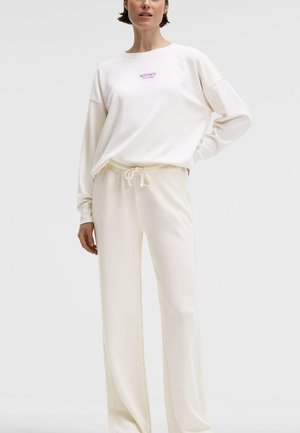 Frau trägt cremefarbene Jogginghose und passendes langärmeliges Sweatshirt mit lila "Senses The Label" Logo, steht vor einem einfarbigen Hintergrund.