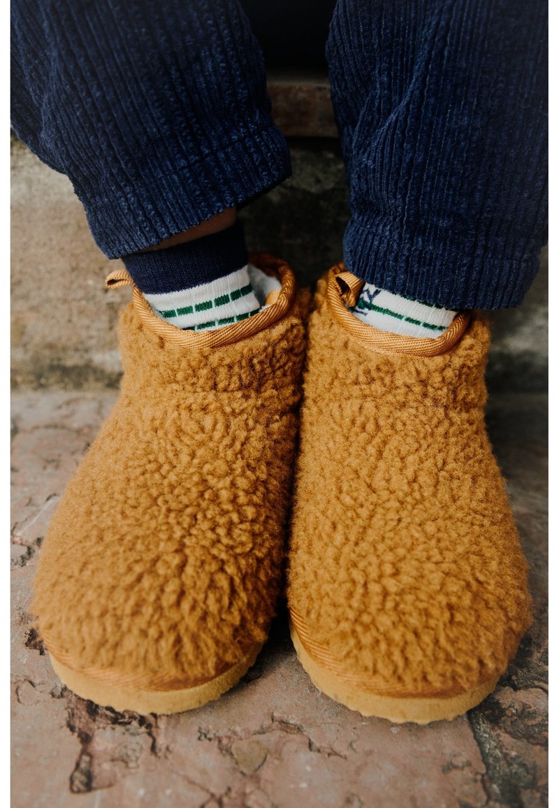 Pieds portant des chaussons duveteux orange-brun, des chaussettes blanches avec des rayures vertes et un pantalon en velours côtelé bleu foncé, debout sur un sol en pierre texturée.