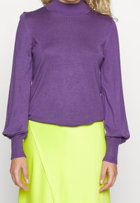 Fialový top s dlouhým rukávem a vysokým límcem, s žebrovanými manžetami, spárovaný s jasně žlutou zavinovací sukní. Oba kousky mají hladké, jemné textury.