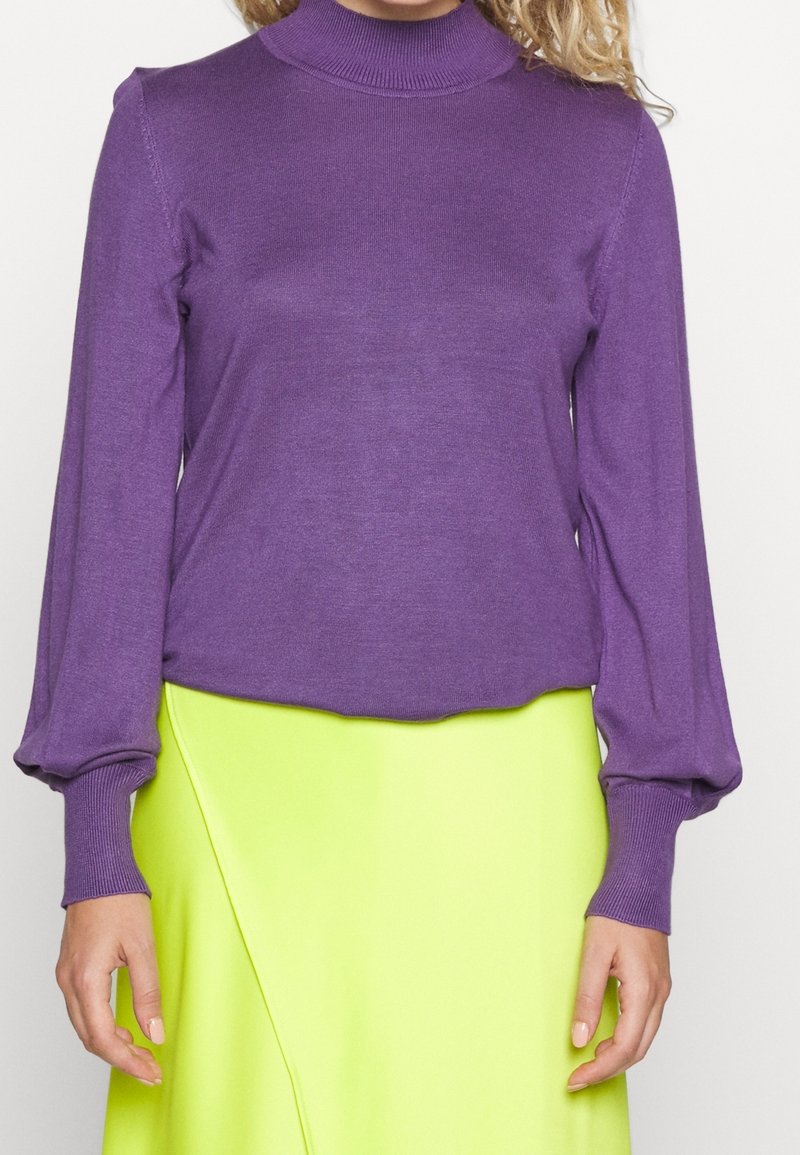Fialový top s dlouhým rukávem a vysokým límcem, s žebrovanými manžetami, spárovaný s jasně žlutou zavinovací sukní. Oba kousky mají hladké, jemné textury.