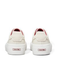 Calvin Klein sko har en crème farvet canvas-overdel, bølget design, røde detaljer nær hælen og en hvid gummisål med logo-detalje.