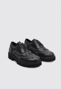 Schwarze Leder Schuhe mit Broguemuster, silbernen Nietenakzenten, runder Zehenpartie und dicker schwarzer Gummisohle für zusätzlichen Komfort.