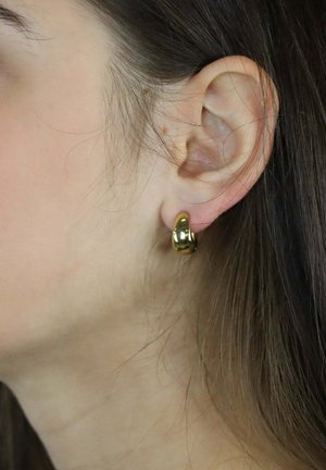 Ronde oorring in goudkleur, gedragen aan een oor, met een gladde, glanzende oppervlakte en een eenvoudig, modern ontwerp.