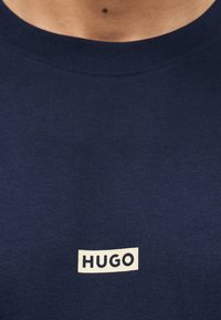 Μπλουζάκι ναυτικό μπλε με στρογγυλή λαιμόκοψη και μικρό λευκό ορθογώνιο λογότυπο "HUGO" στο κέντρο του στήθους κάτω από τη λαιμόκοψη.