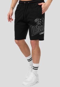 Hombre vistiendo pantalones cortos negros de Lonsdale London, calcetines deportivos blancos con rayas negras y zapatillas blancas, de pie contra un fondo gris.