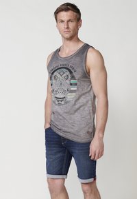 Siva majica bez rukavov z grafiko lobanje in geometrijskimi vzorci ter modrimi akcenti, v kombinaciji s temnimi denim kratkimi hlačami s zvitimi robovi.