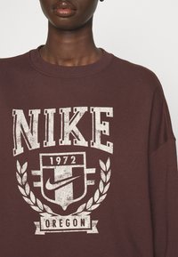 Vinröd sweatshirt med en vit sliten logotyp, texten "NIKE" och "OREGON", med ett sigill och lagerbladsmotiv. Mjuk bomullsblandning.