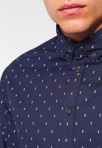 Marineblauw button-up shirt met een kraag, kleine witte ruitjespatroon en zwarte knopen. Glad materiaal met een getailleerd ontwerp.