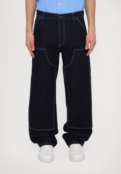 Pantaloni din denim de culoare albastră închisă, cu picioare largi, cusături contrastante de culoare albastră deschisă, buzunare frontale și închidere cu nasture. Purtați cu adidași albi.