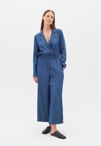 Denim salokleita ar dziļu V veida kakla izgriezumu, garām piedurknēm un elastīgu jostasvietu. Plašas kājas, gaišā mazgāšanā un redzamām šuvēm. Melni slīdējošie apavi.