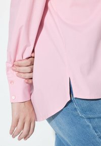 Camicia rosa chiaro con taglio dritto, polsini con bottoni, sottili spacchi laterali, indossata sopra jeans in denim, tessuto di cotone liscio.