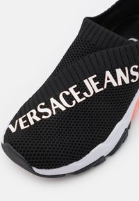 Sort slip-on sneaker med tekstureret strikket overdel og stor "VERSACE JEANS" tekst på toppen, med hvide og sorte såls detaljer.