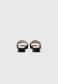 Black slip-on shoes made of suede, featuring a shiny black heel, gold studs on the straps, and a smooth beige insole.