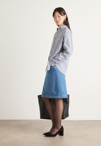 Randig skjorta med knapp, blå denimkjol som går till knäna, svarta klackar, håller en svart tote-väska, står på en neutral golvbakgrund.