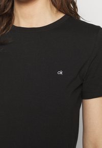 Kobieta nosząca gładką, czarną koszulkę z krótkim rękawem z małym, białym haftowanym logo "ck" na lewej piersi.