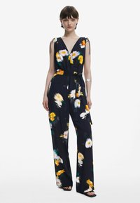 Blommig jumpsuit med djup V-ringning, marinblå bakgrund, gula och vita blommor, justerbara bindremmar och ett matchande tygbälte i midjan.