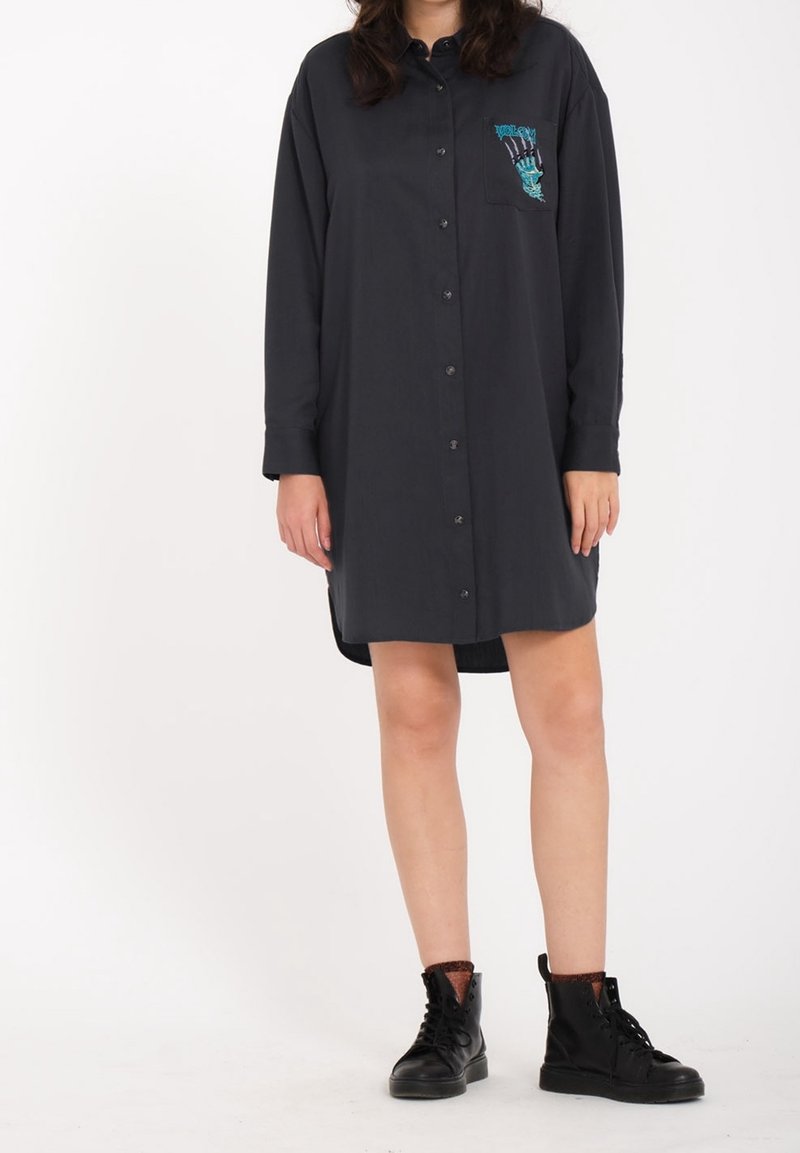 Robe noire à manches longues avec un bouton et une poche poitrine, ornée d'un graphique de main turquoise. Associée à des bottines noires au niveau de la cheville. Texture de tissu lisse.