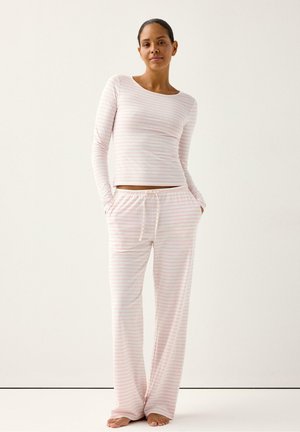 Kvinde står barfodet iført lyserød og hvidstribet langærmet top og matchende bukser med snøre foran mod ensfarvet baggrund.