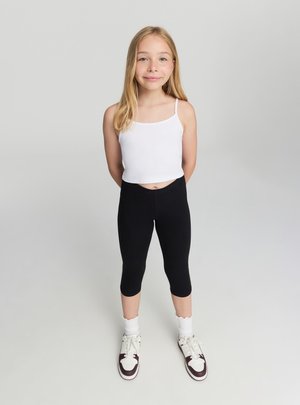 Ragazza giovane in piedi con le mani dietro la schiena, indossa una canotta bianca, leggings capri neri, calzini bianchi e scarpe da ginnastica bianche con dettagli marroni.