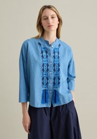 Camicia in denim azzurro chiaro con bottoni, decorata con motivi blu ricamati e nappine all'orlo. Presenta un colletto alla cinese e maniche tre quarti.