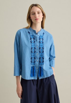 Light blue denim button-up shirt with blue embroidered patterns and tassels at the hem. Features a mandarin collar and three-quarter sleeves.