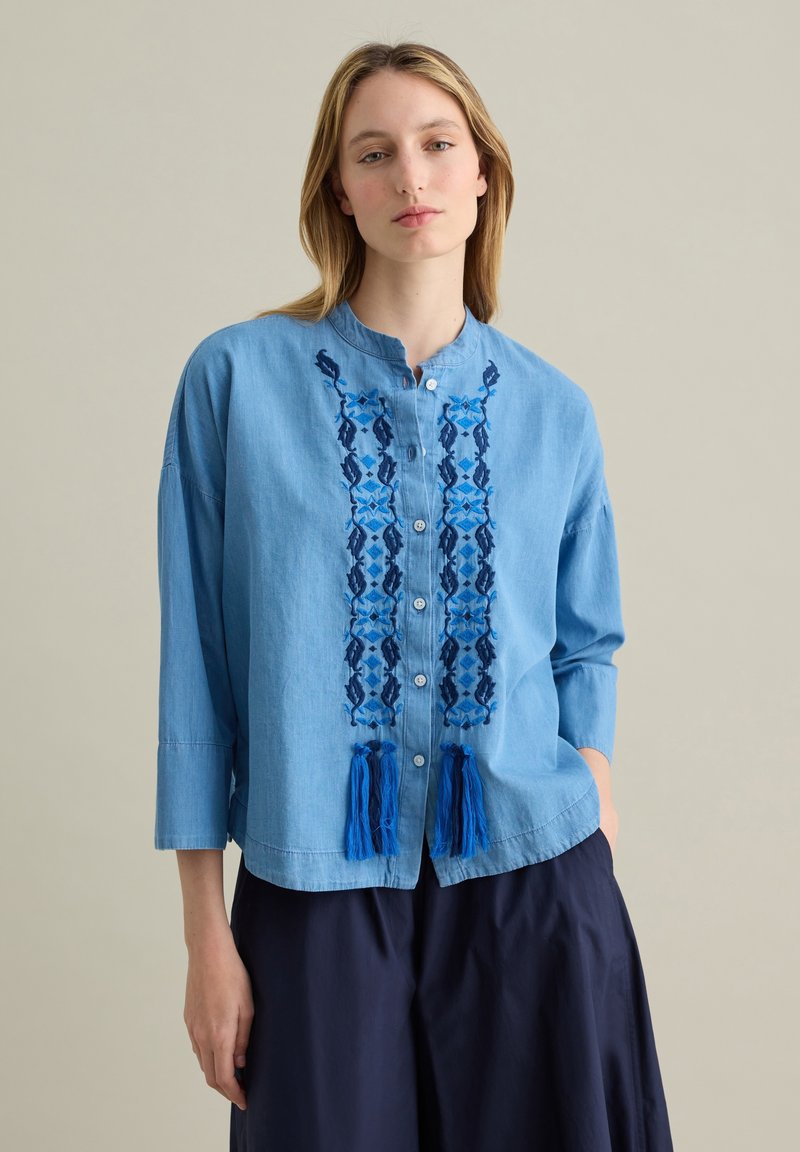Lichtblauwe denim overhemd met knopen, versierd met blauwe geborduurde patronen en franjes aan de zoom. Beschikt over een mandarinakraag en driekwart mouwen.