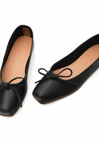 Bailarinas negras de cuero con punta redonda, que presentan un lazo delgado en la parte frontal y un interior suave. La suela es de color beige claro.