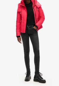 Różowa kurtka puchowa z zamkiem błyskawicznym i regulowanym kapturem, w połączeniu z czarnym swetrem, ciemnymi obcisłymi dżinsami i czarnymi sznurowanymi botkami za kostkę.