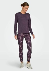 Langærmet lilla skjorte kombineret med mønstrede leggins med mørke og lyse abstrakte designs. Model iført lysegrå sneakers.