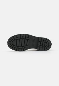 Semelle de chaussure en caoutchouc noir avec un motif de semelle texturé, présentant de profondes rainures et des grips circulaires surélevés pour une meilleure traction.