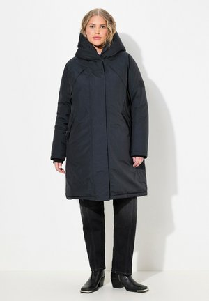Abrigo negro aislante con cuello alto, mangas largas y bolsillos laterales, llevado sobre jeans oscuros y botas de tobillo. Textura suave, con un minimalismo en los detalles.