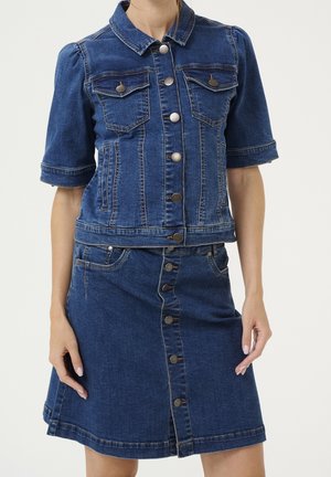 Femme portant une veste courte en denim bleu à manches courtes et une jupe en denim en ligne A avec boutons sur le devant, debout devant un fond uni.