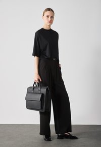 Top negro de manga corta, pantalones anchos negros y zapatos negros pulidos. Se sostiene un bolso de piel negra estructurado con un asa superior.